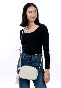 Kvinde iført sort lang ærmet top og blå jeans med en hvid skuldertaske med kvast, der ser til siden mod en hvid baggrund.