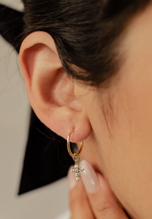 Gold hoop earring with a small cross pendant, adorned with clear gemstones, hanging near the ear. Smooth texture and polished finish.