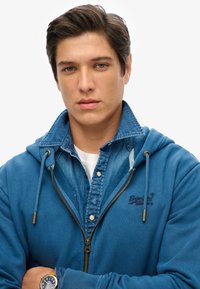 Sudadera azul con cremallera colocada sobre una camisa de mezclilla, que cuenta con capucha ajustable, logo bordado y cierre con cremallera. Tejido texturizado.