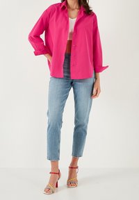 Camicia fucsia con bottoni e maniche arrotolate, abbinata a jeans cropped blu chiaro e sandali colorati con tacco alto.