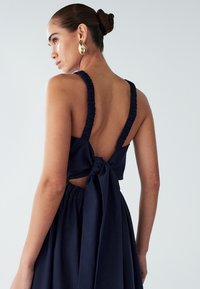 Kvinna med håret i en knut iförd en marinblå ärmlös klänning med draperade axelband och en stor rosett knuten vid midjan, mot en enfärgad bakgrund.