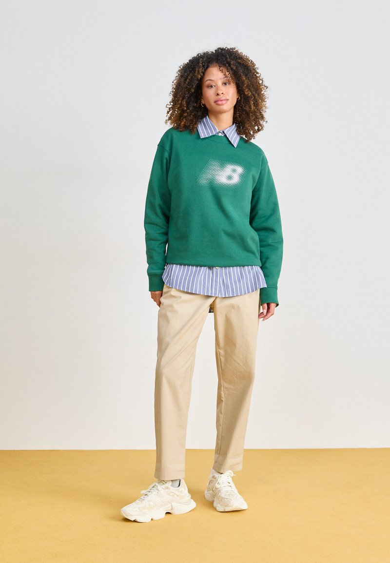 Sudadera verde con un logotipo gráfico, superpuesta a una camisa de rayas azul y blanca, combinada con pantalones beige y zapatillas blancas.