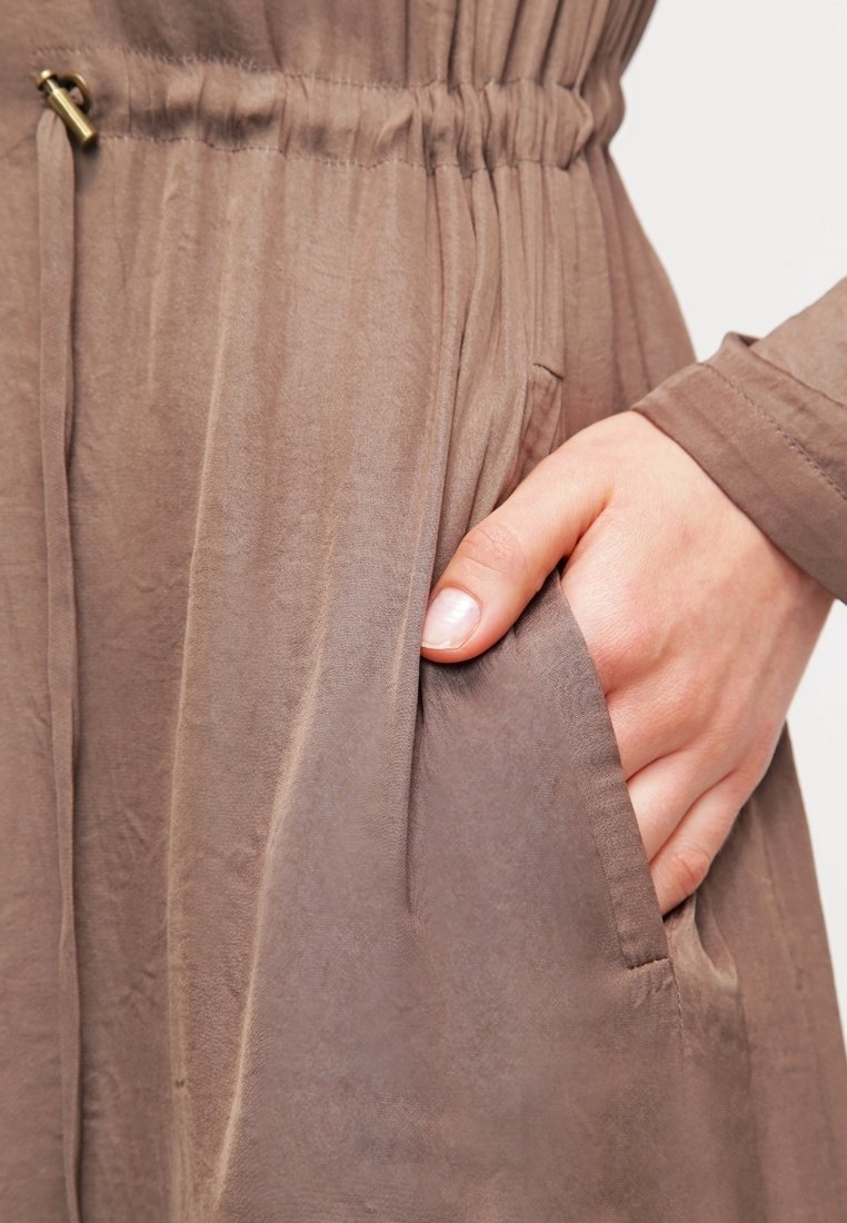 Hellbraunes Kleid aus glattem, leichtem Stoff. Verfügt über einen elastischen Bund und eine Seitentasche, in der eine Hand ruht.