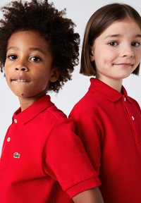 Due bambini indossano polo rosse con una finitura strutturata. Le magliette presentano un colletto a tre bottoni e il logo Lacoste sul lato sinistro.
