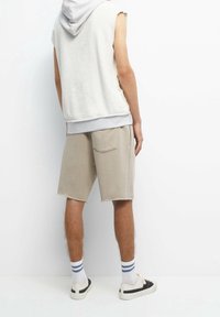 Light gray sleeveless hoodie with terry texture, paired with light beige shorts featuring a back pocket. White and black sneakers with striped socks.
