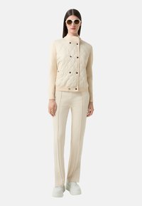 Jasno beżowa kurtka pikowana o dopasowanym kroju, czarne guziki i kremowe rękawy, w połączeniu z dopasowanymi kremowymi spodniami i białymi sneakersami na platformie.