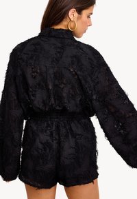 Femme portant un combishort noir texturé à manches longues avec une taille élastique, des boucles d'oreilles en forme de cerceau dorées, de dos sur un fond clair.