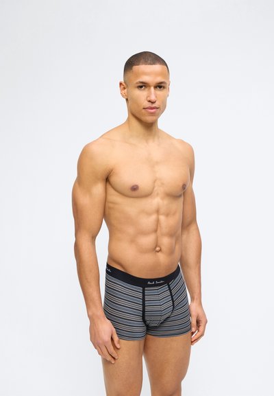 Μυώδης άνδρας στέκεται, φορώντας γκρι ριγέ boxer briefs με μαύρη μέση, μπροστά σε ένα απλό φωτεινό φόντο.