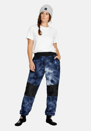 Frau in weißem T-Shirt, grauer Mütze und blauen Jogginghosen mit Batikmuster und schwarzen Knieschonern, die mit den Händen in den Taschen steht.