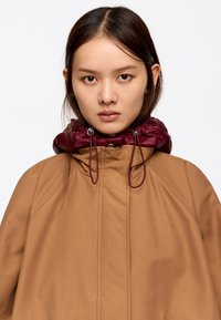 Brązowa wodoodporna kurtka z bordowym satynowym kołnierzem, z regulowanymi sznurkami i zapięciem na napy. Gładka tekstura i luźny krój.