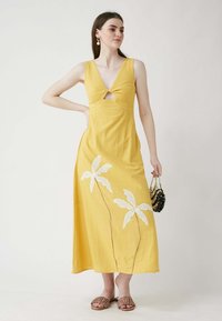 Gelbes Baumwoll-Maxikleid mit tiefem V-Ausschnitt, Knoten-Detail vorne und weißer Blumenstickerei am Saum. Abgerundet mit einer schwarzen Tasche.
