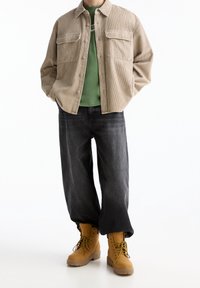 Chaqueta de pana beige con dos bolsillos frontales, camiseta verde, jeans de pierna ancha gris oscuro y botas marrones con cordones y suelas con patrón.