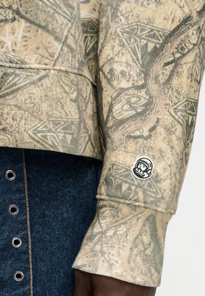 Μπεζ φούτερ με υφασμάτινο σχέδιο δέντρων και μαύρο λογότυπο στην μανσέτα. Τζιν φούστα με ασημένια κουμπιά snap εμφανή.