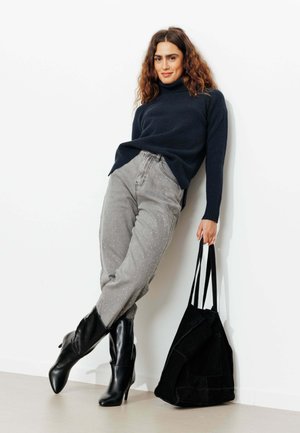 Pull marin bleu, pantalon en denim gris avec des accents cloutés argentés, bottes à talons noirs, et un grand sac fourre-tout noir.