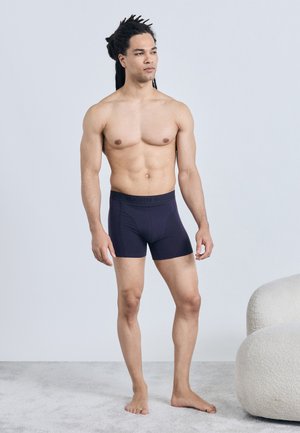 Uomo senza maglietta con dreadlocks che indossa boxer blu navy in piedi su un tappeto vicino a una sedia beige curva contro una parete chiara semplice.