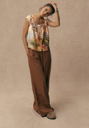 Short-sleeve floral blouse with gathered neckline, paired with loose brown trousers featuring paisley embroidery and tassel-accented footwear.