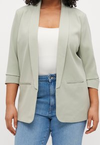 Lichtgroene blazer met een gestructureerd ontwerp, open voorkant en ¾ mouwen; gecombineerd met hoge taille blauwe jeans. Gladde stof, minimalistische accenten.