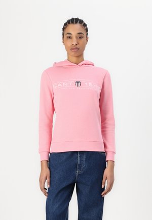 Pink hoodie made of soft cotton fabric, featuring a front logo print, adjustable drawstring hood, and ribbed cuffs and hem. Worn with denim.