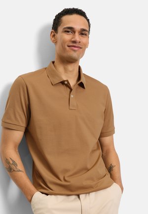 Homme souriant aux cheveux courts et bouclés, portant un polo marron et un pantalon beige, debout les mains dans les poches devant un fond blanc.
