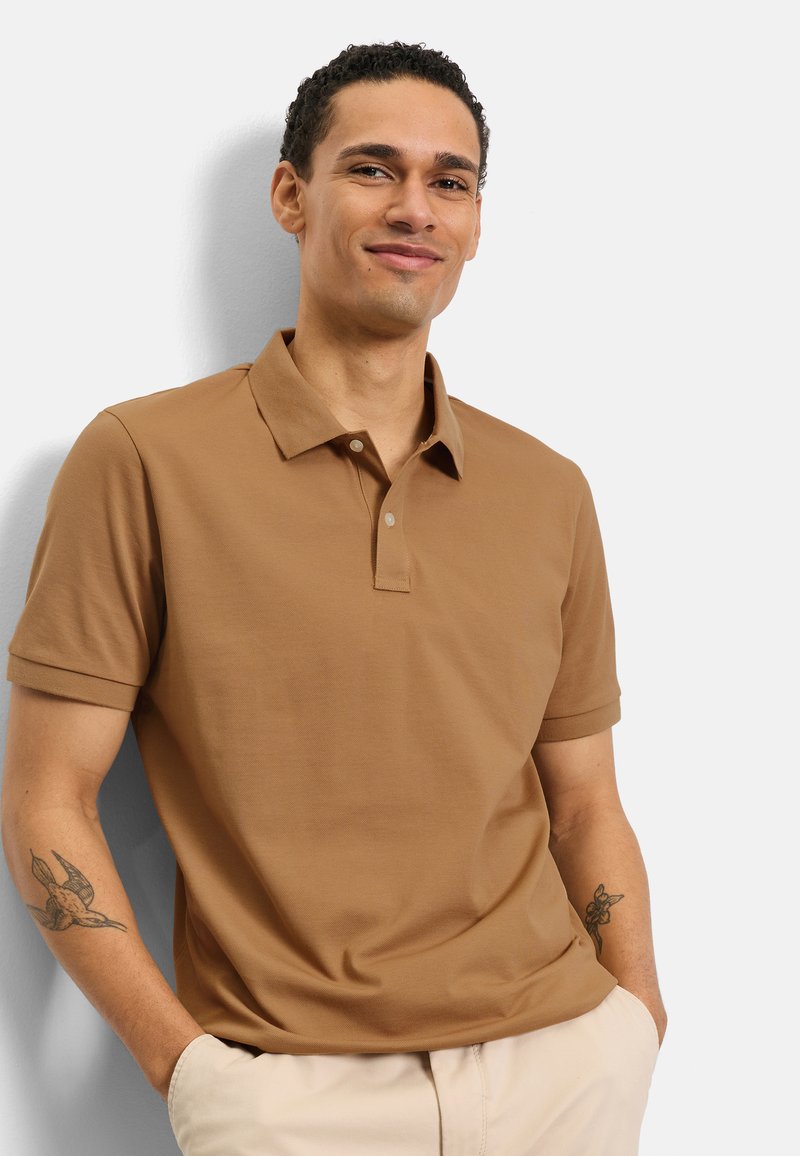 Uomo sorridente con capelli corti e ricci che indossa una polo marrone e pantaloni beige, in piedi con le mani in tasca davanti a uno sfondo bianco.