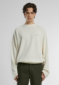 Hellecremefarbene Langarmpullover mit strukturiertem Muster, lockerer Passform, geripptem Kragen und subtil gesticktem Logo auf der Brust.
