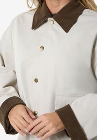 Kvinde iført en lyse beige jakke med brune fløjlskraver og manchetter, bronze trykknapper og forlommer, hænderne samlet ved livet.