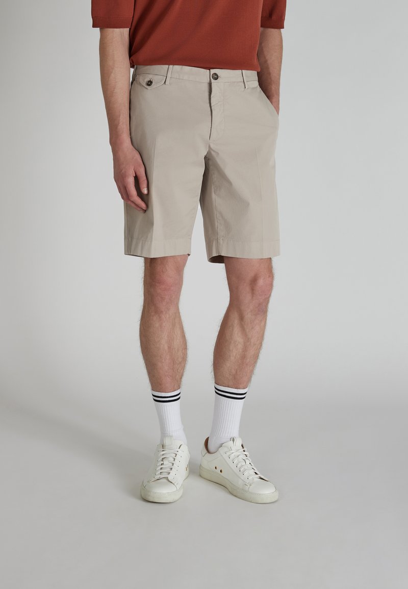  bermuda in cotone beige con frontale piatto, due tasche anteriori e chiusura a bottone. Indossati con sneakers bianche e calzini a righe a costine.