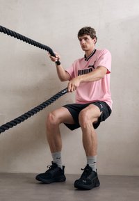 Sportaš u ružičastoj sportskoj majici s crnim detaljima, crnim kratkim hlačama i crnim tenisicama, koristi crne ratne uže na neutralnoj pozadini.