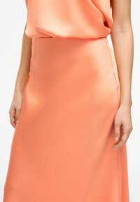 Jupe longue en satin de corail solide avec une texture lisse, détail de couture latérale et un design fluide. Main positionnée sur le vêtement.