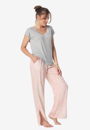 Kobieta stojąca boso, ubrana w luźną szarą bluzkę z dekoltem w serek oraz szerokie różowe wzorzyste spodnie piżamowe z bocznymi rozcięciami i kieszeniami.