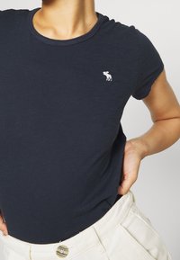 Navy blue cotton t-shirt with short sleeves, ribbed neckline, and small white moose logo on the chest. Worn with white high-waisted pants.