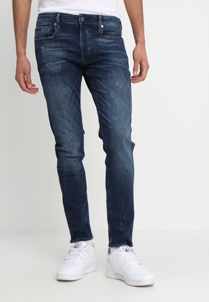Homme portant un jean slim bleu foncé et des baskets blanches, debout devant un fond clair uni.