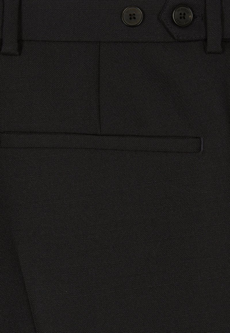 Primer plano de pantalones negros de tela texturizada con cinturilla, cierre de doble botón, trabillas para cinturón y bolsillo con abertura.