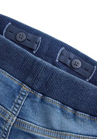 Denimbukser med en blå ribbet taljebånd med to sorte knapper og kontrastfuld gul syning langs sømmene.