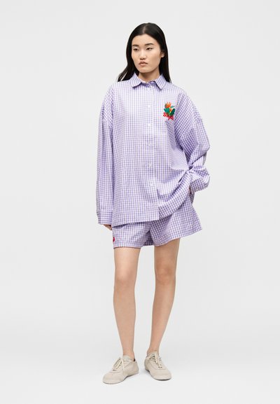 Νεαρή γυναίκα φοράει υπερβολικά μεγάλο πουκάμισο λευκό με μοβ καρό και ταιριαστά σορτς με λευκά αθλητικά, στέκεται μπροστά σε φωτεινό μονόχρωμο φόντο.