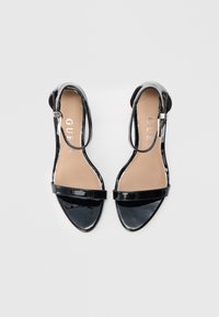 Sandalias de charol negro con un diseño elegante, punta afilada y correa en el tobillo. Cuentan con un tacón plano y una plantilla lisa de color beige.