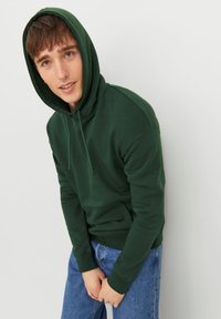Felpa verde in cotone con tasca frontale, cappuccio regolabile con cordino e polsini a coste, abbinata a jeans in denim blu chiaro.