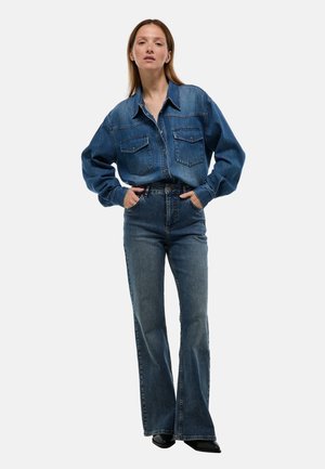 Chemise en denim avec deux poches poitrine, de couleur bleue, coupe décontractée. Assortie à un jean flare taille haute en denim bleu assorti, avec une ceinture.