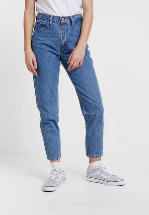 Person wearing high-waist blue jeans, white socks, gray and white sneakers, and a tucked-in white t-shirt, standing on a plain background.