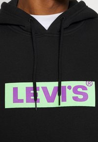Černá mikina s kapucí s zeleným obdélníkovým logem s nápisem "LEVI'S®" tučným fialovým písmem. Obsahuje nastavitelné šňůrky a má měkkou texturu.