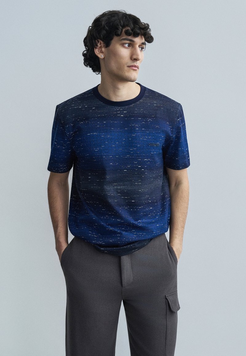 Navy-blue short-sleeve t-shirt with a textured pattern, round neck, and subtle branding on the chest, paired with gray trousers.