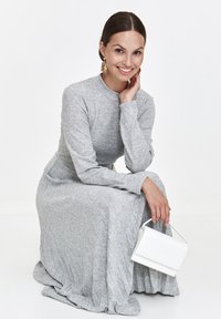 Eine Frau, die in einem hellgrauen, langärmeligen Kleid kniet, lächelt, trägt goldene Ohrringe und hält eine kleine weiße Handtasche vor einem schlichten weißen Hintergrund.