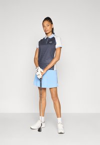 Tumši zila un balta polo krekls ar teksturētu rakstu, īsām piedurknēm un gaiši zilu svārku. Valkāts kopā ar golfa nūju un baltām cimdu.