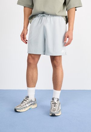 Mężczyzna ubrany w jasnoszorty Nike, białe skarpetki do kostek i szare buty sportowe Nike, stojący na niebieskiej podłodze na tle białej ściany.