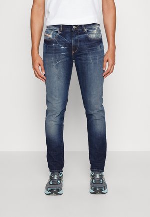 Homme portant un jean slim bleu foncé délavé et des baskets grises avec des détails bleu clair, debout devant un fond clair uni.