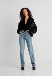 Γυναίκα με σγουρά μαλλιά, φοράει μαύρο κοντό μπουφάν πάνω από μαύρο τοπ, ανοιχτό μπλε τζιν ψηλόμεσο, μαύρη ζώνη και μαύρες μυτερές μπότες, στέκεται όρθια.