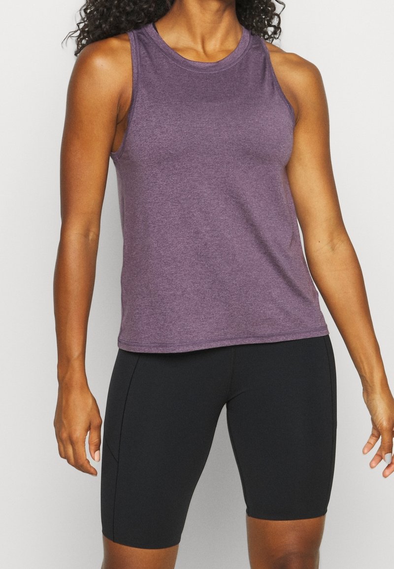 Personne athlétique portant un haut violet sans manches et un short de cyclisme noir moulant, debout devant un fond uni.