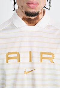 White polo shirt with light gold horizontal stripes. Features bold gold letters "A I R" and a small gold Nike logo. Smooth texture.