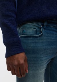 Ναυτικό πλεκτό πουλόβερ με ριμπ μανσέτες, συνδυασμένο με ξεβαμμένο μπλε τζιν παντελόνι που διαθέτει μια μικρή τυρκουάζ λεπτομέρεια στην πίσω τσέπη.