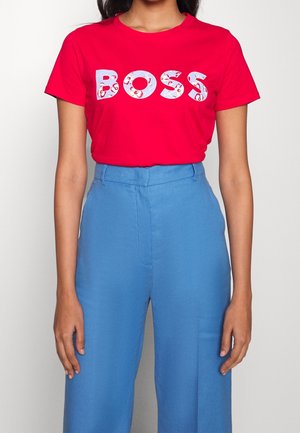 Kobieta w czerwonym T-shircie z napisem "BOSS" w ozdobnych literach i jasnoniebieskich, wysokich spodniach na tle jednolitego tła.